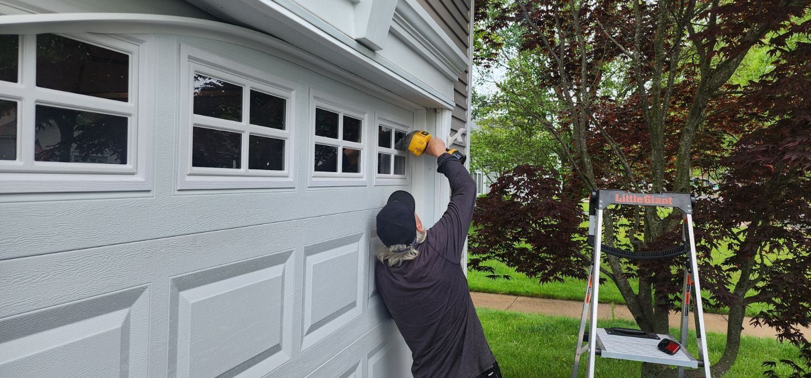 A person wearing a dark shirt and hat paints the trim of a white garage door while standing on a ladder.