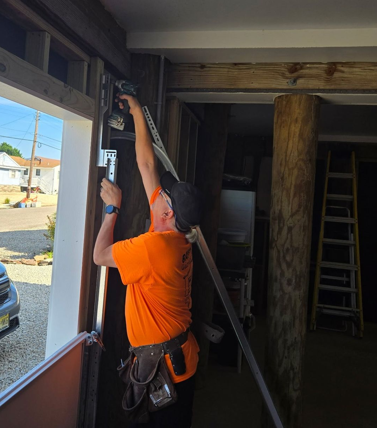 A person wearing an orange shirt and a tool belt uses a power drill to install a metal track on a door frame.