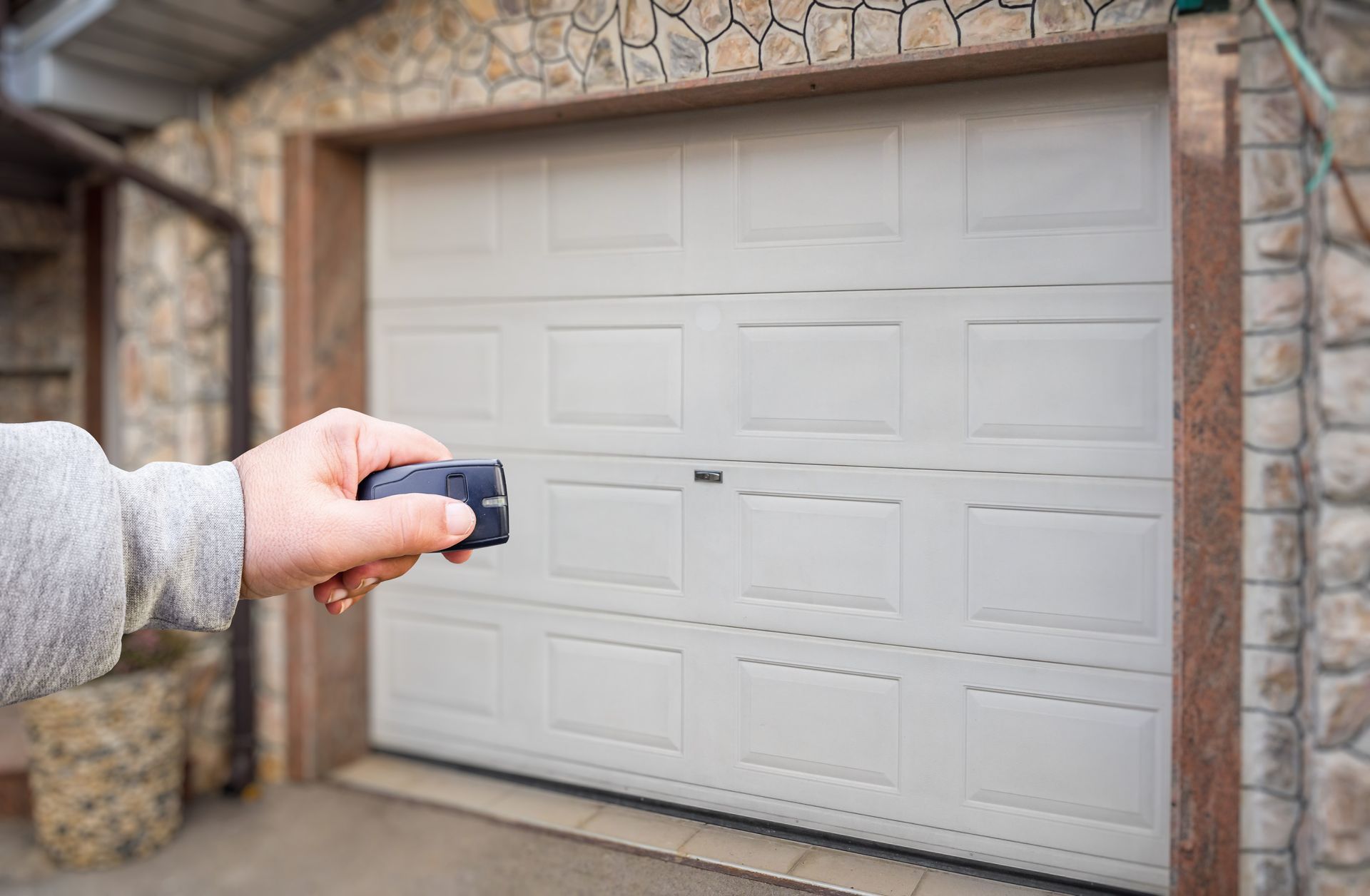 A hand holds a remote control, aiming it at a closed, light-colored panel garage door on a stone building.