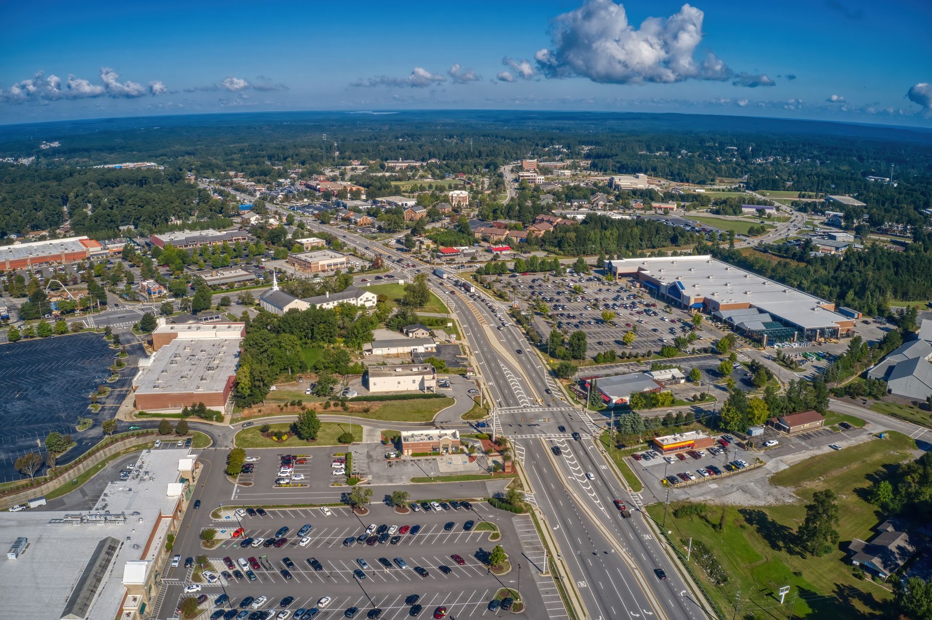 Aerial view of a town with buildings, roads, parking lots, and a forest in the background on a sunny day