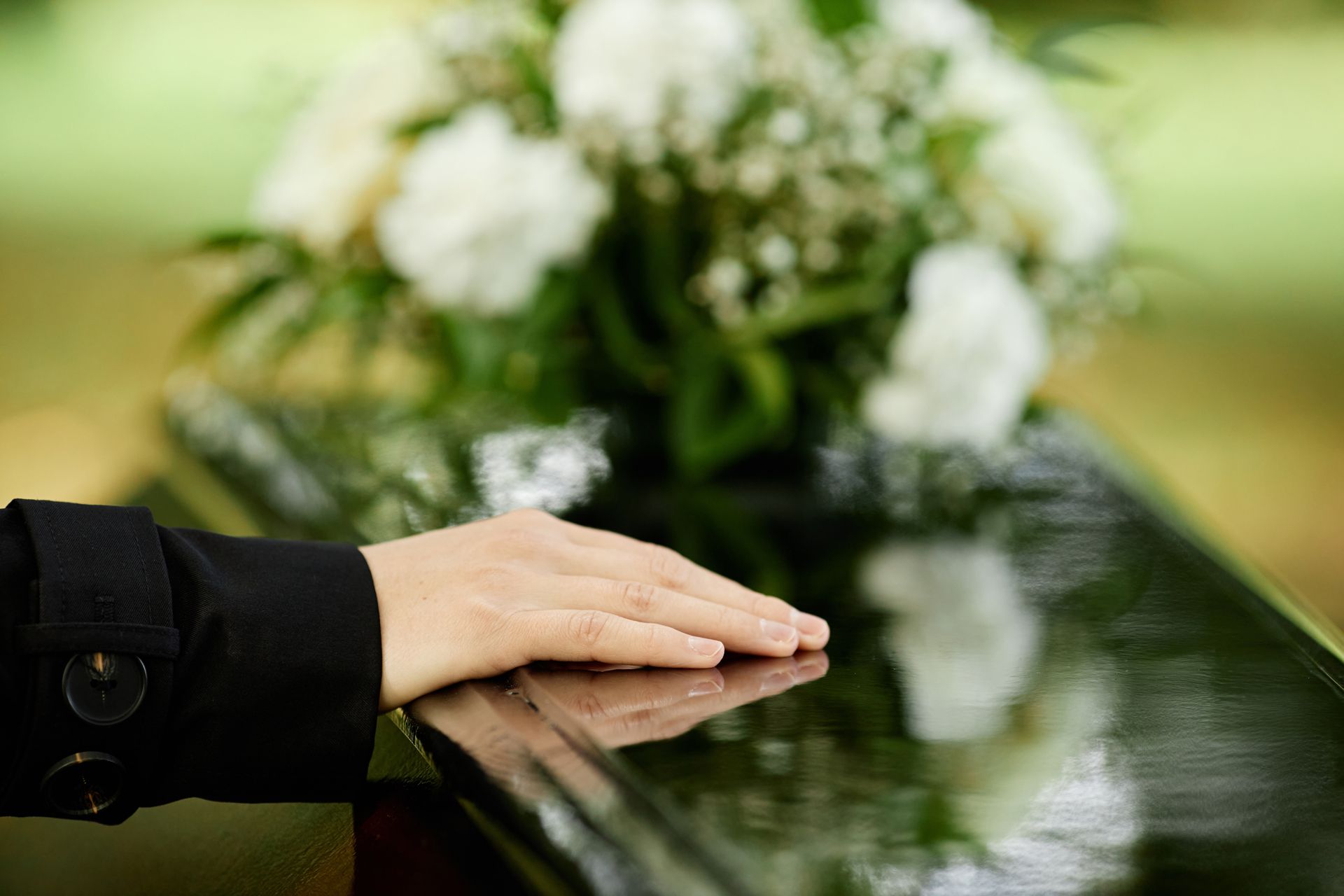Hand resting on a dark casket; white flowers in the background.