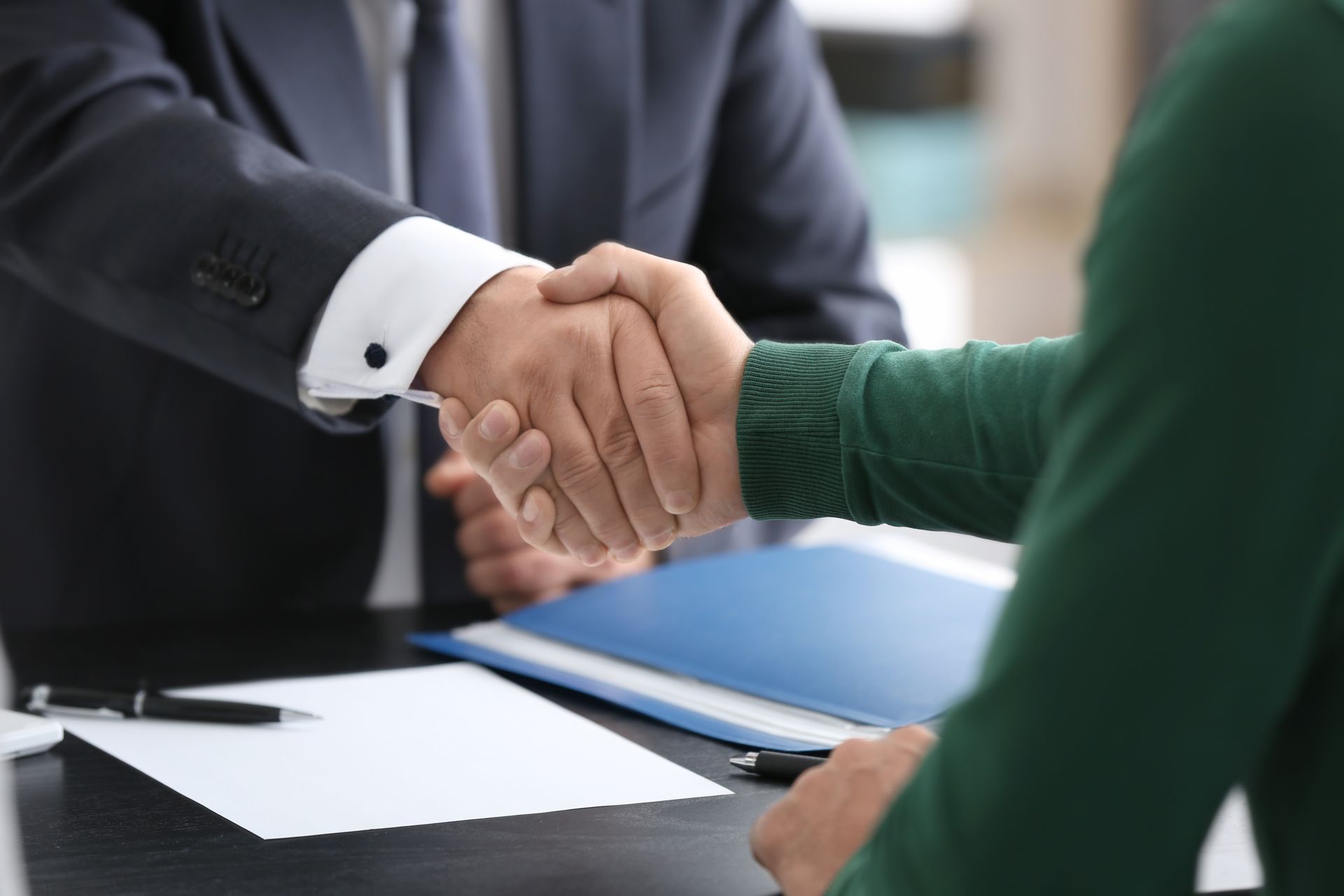 Two people in business attire shake hands over a desk with paperwork.