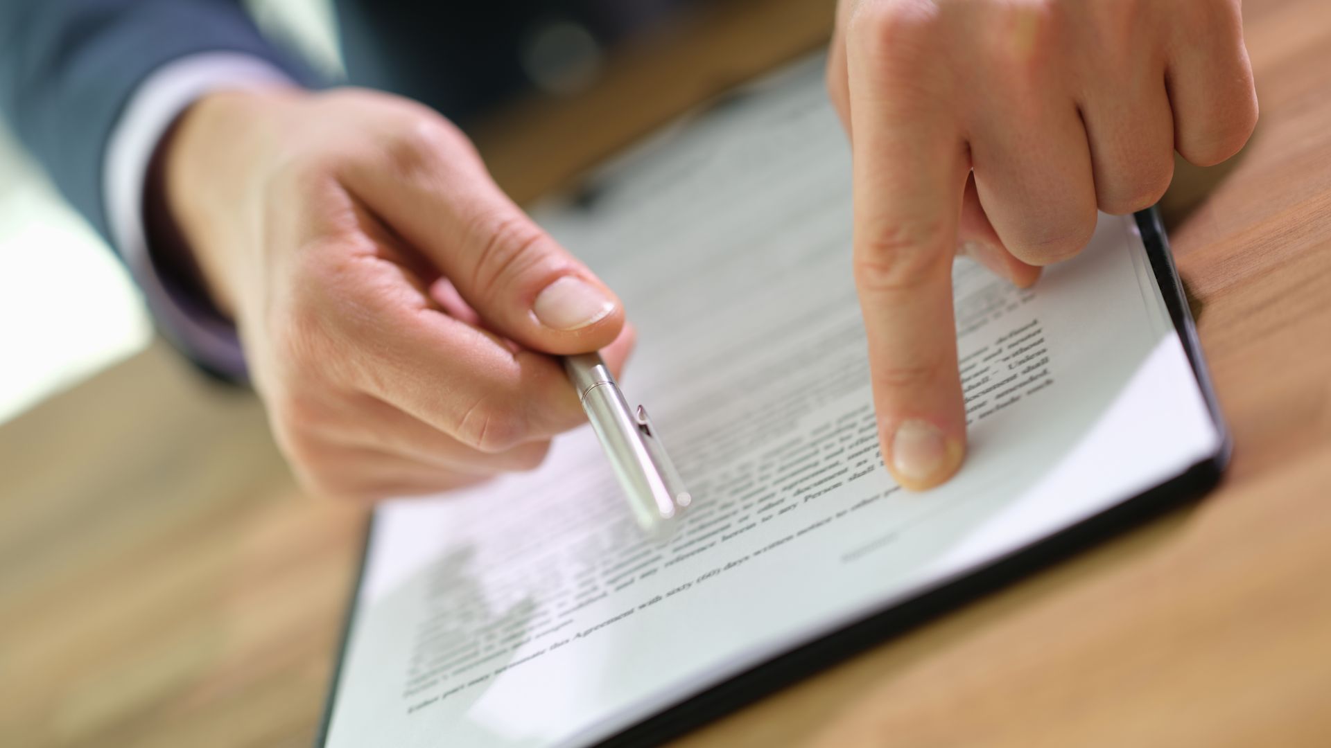 Person's hands pointing at text on a clipboard with a pen on a desk.