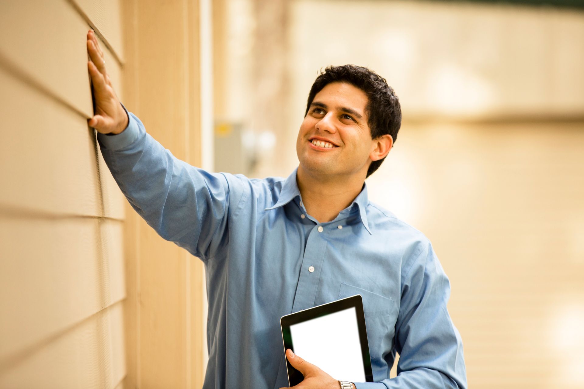 Man in blue shirt, holding tablet, inspecting house siding, smiling, outside.