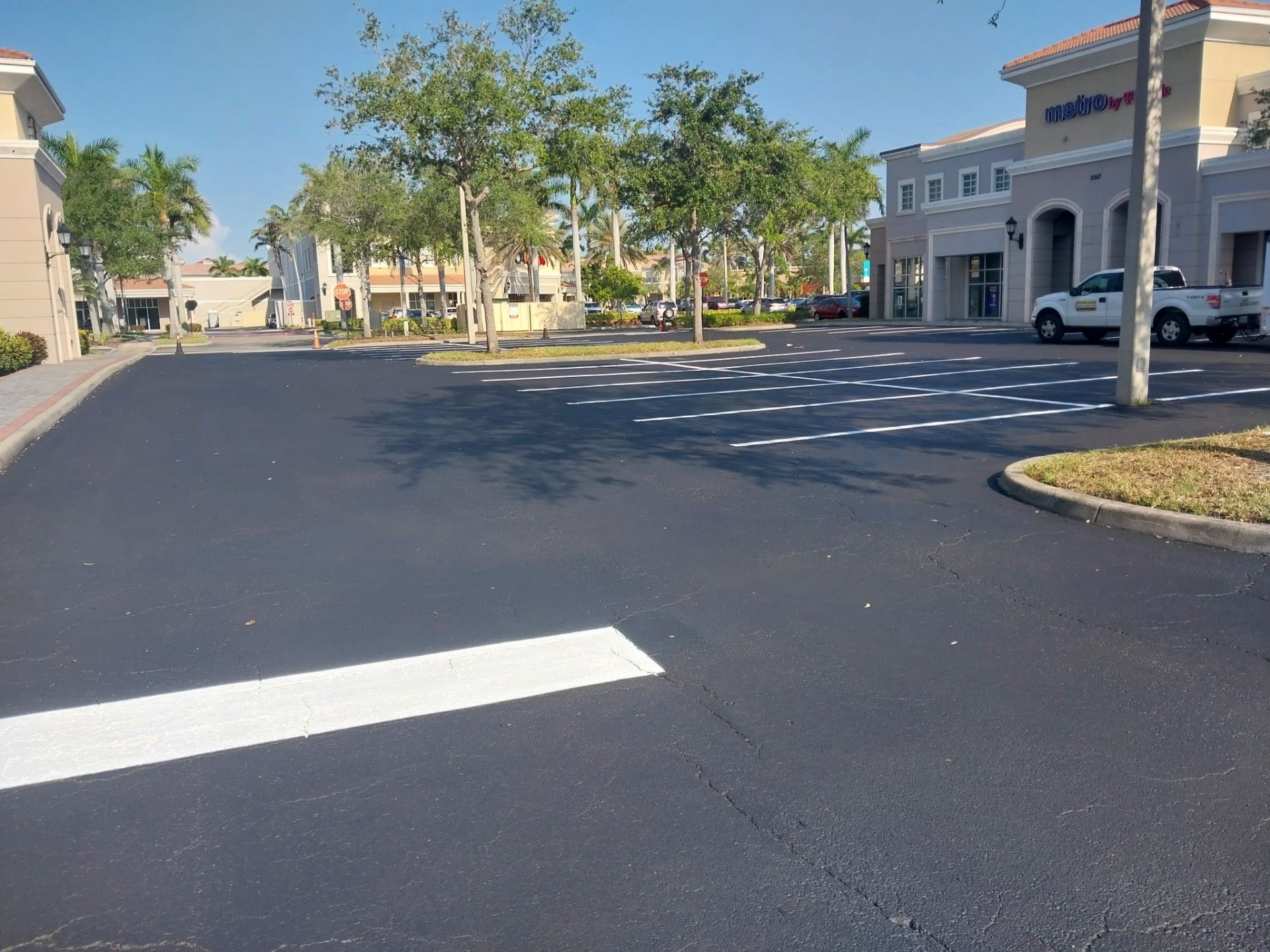 Empty asphalt parking lot with painted crosswalk lines; businesses and trees in the background on a sunny day.