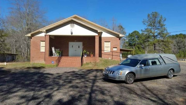 Silver hearse parked in front of a brick church on a sunny day.