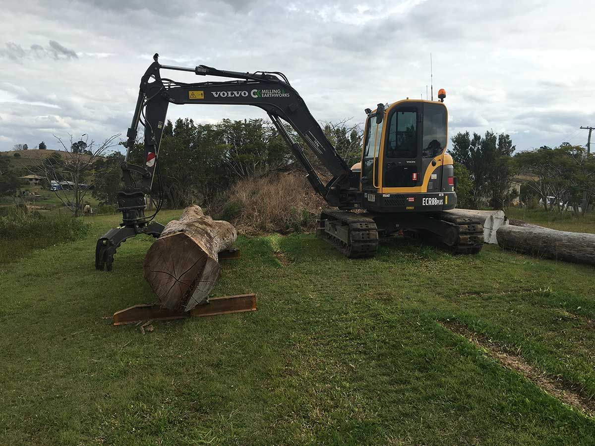 A Large Log Is Being Lifted by An Excavator in A Grassy Field — CK's Milling & Earthworks in Goomeri, QLD