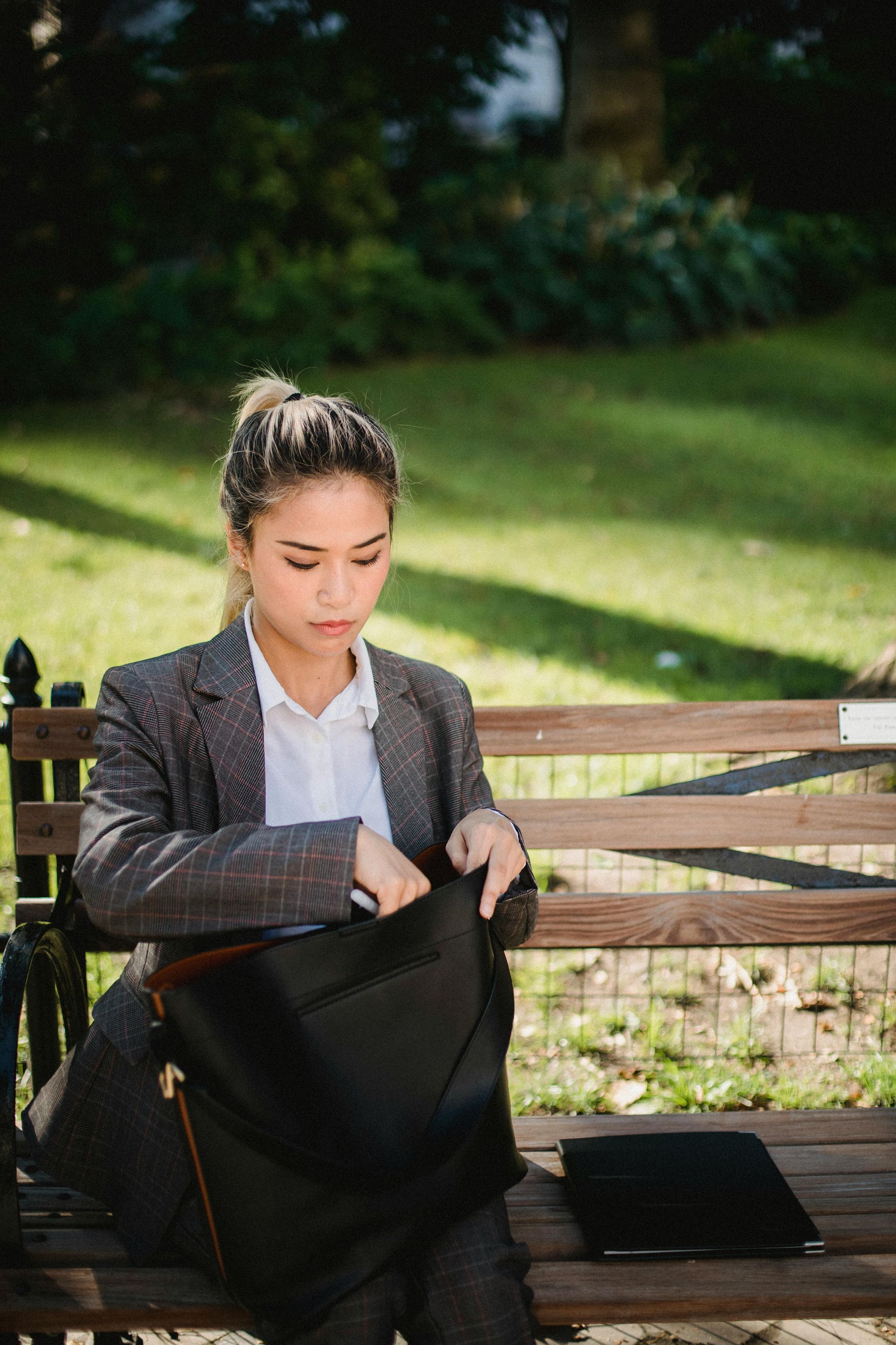 A woman in a suit is sitting on a park bench holding a black bag.