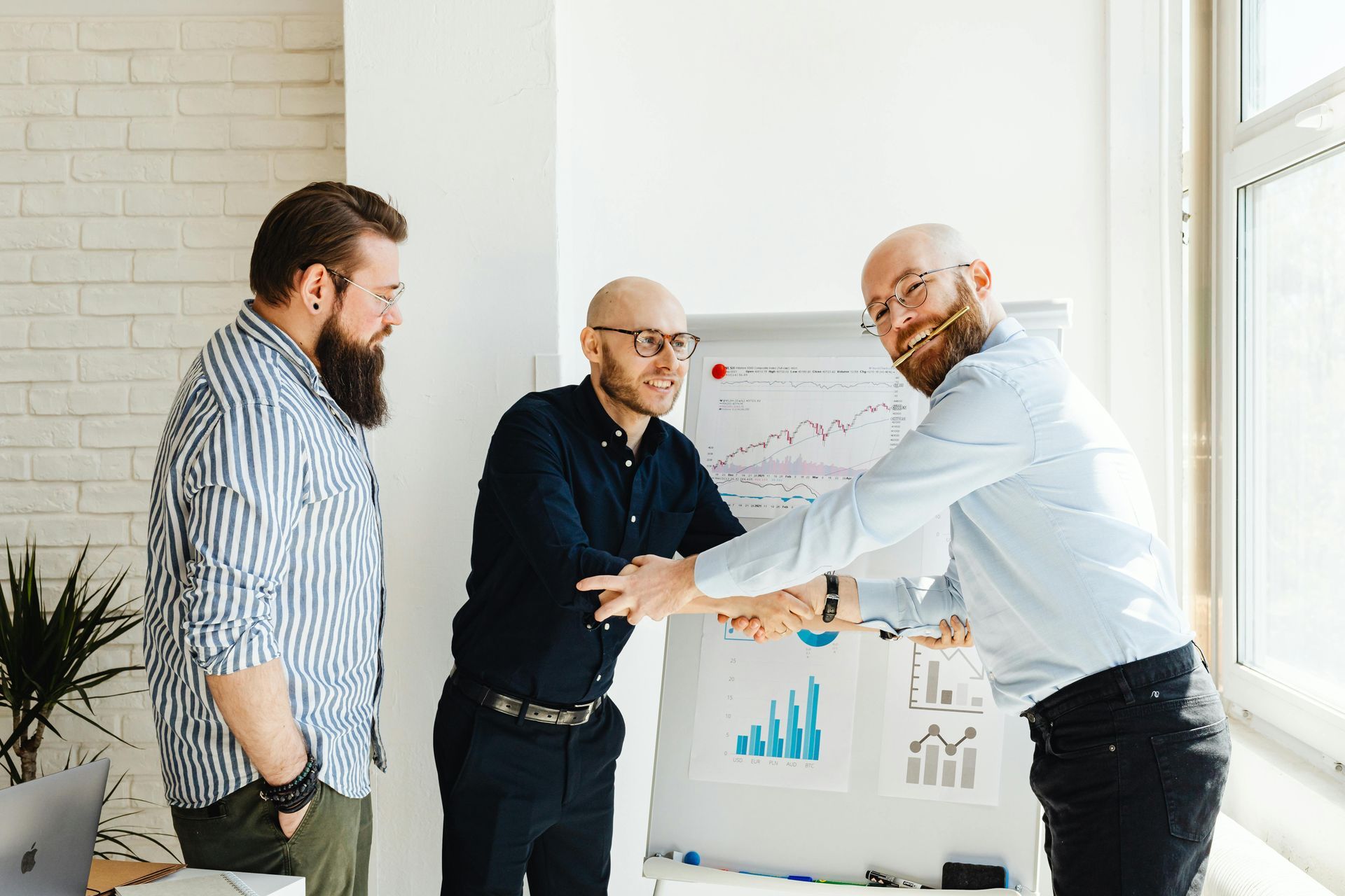 Three men are shaking hands in front of a whiteboard.