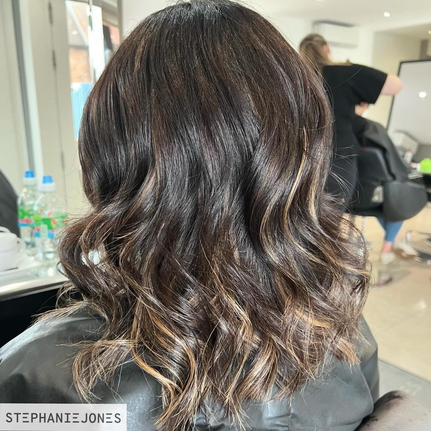 Woman with dark brown hair, styled with waves and blonde highlights, in a salon.
