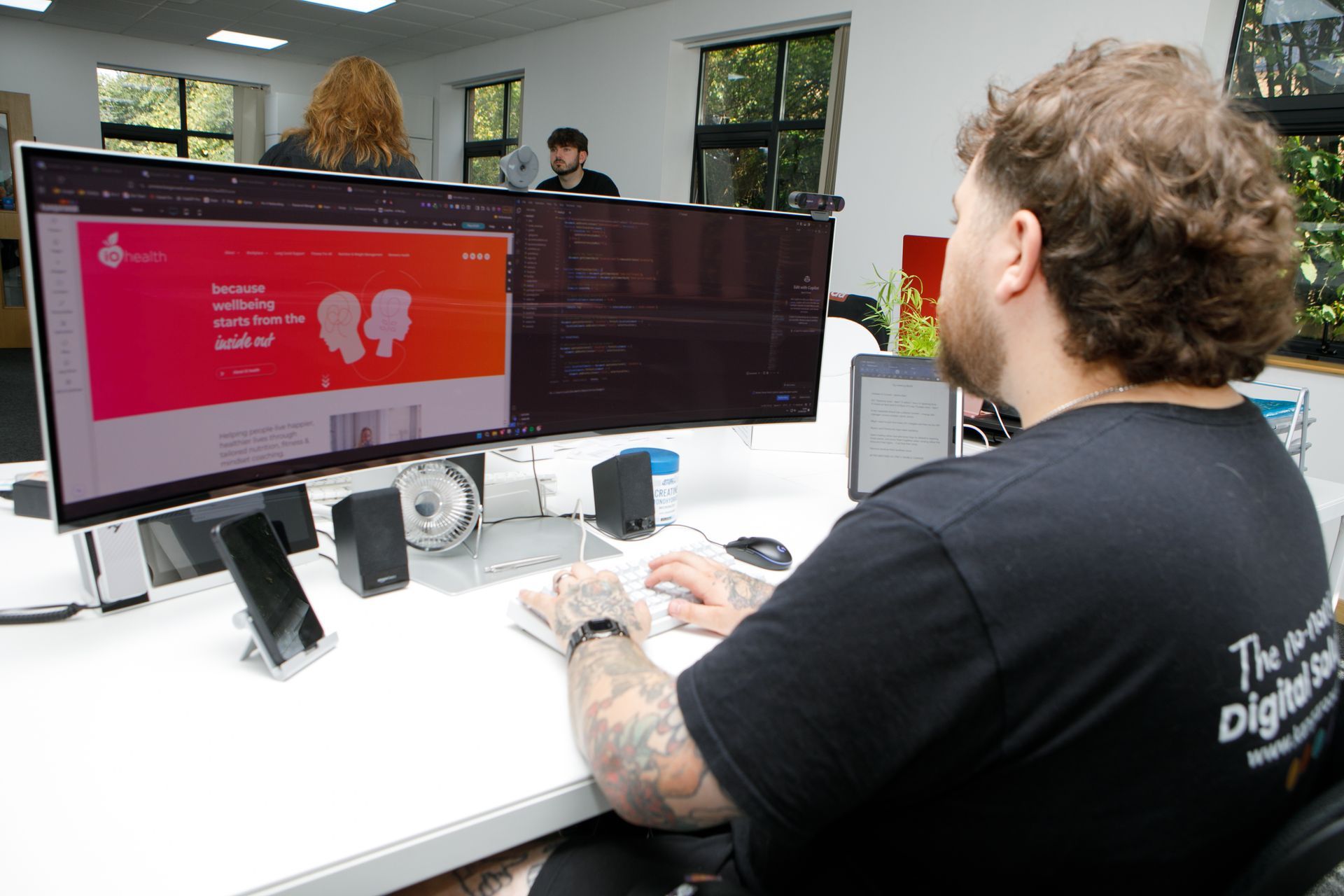 Man with tattoos working on a dual-screen computer, possibly coding, at an office desk.