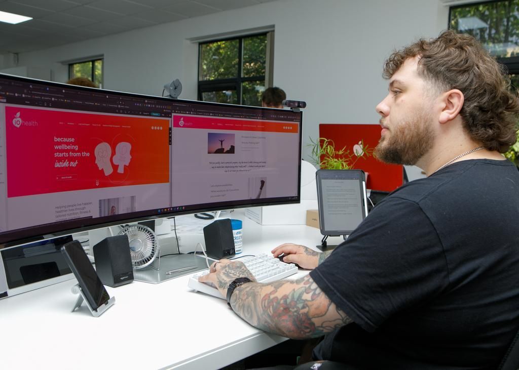 Man with tattoos codes on a computer at a desk, facing three monitors.