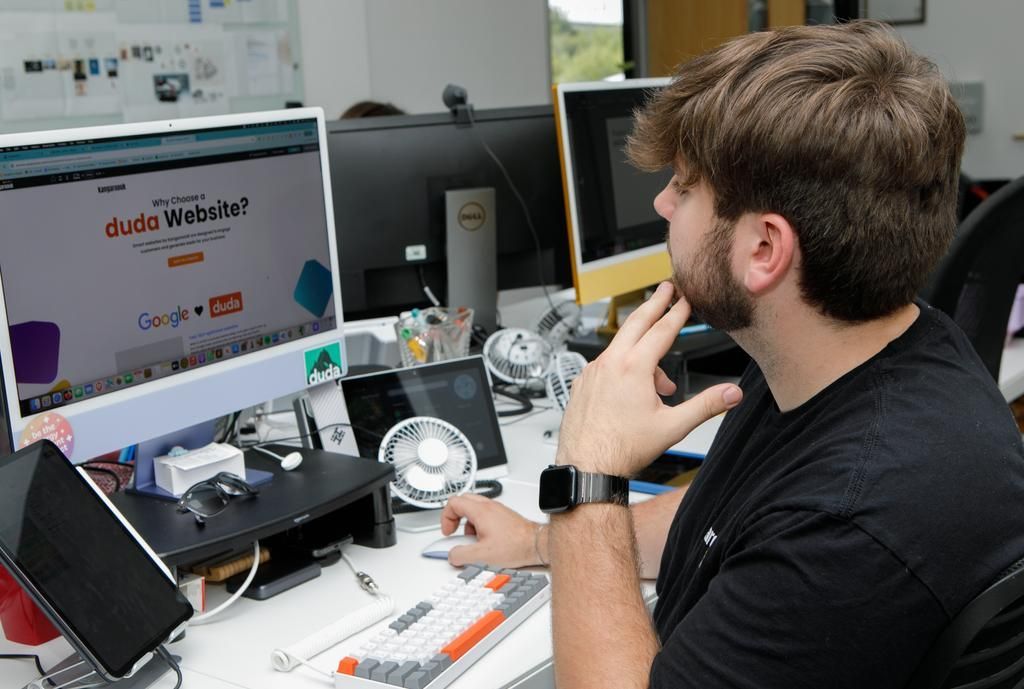 A man at a desk with a computer is pointing at the screen, an office setting.