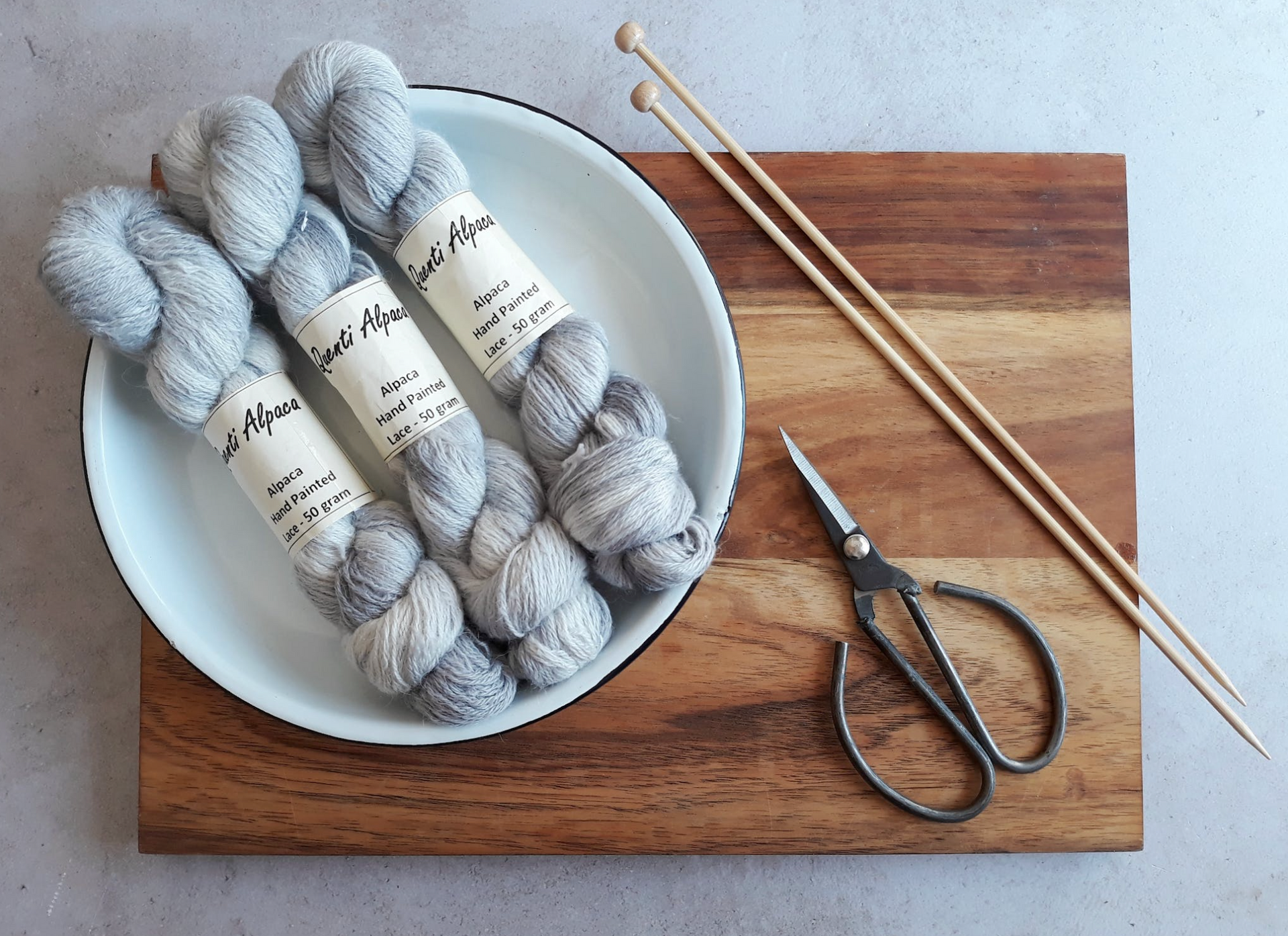 Three skeins of gray speckled yarn in a bowl with knitting needles and scissors on a wooden board.