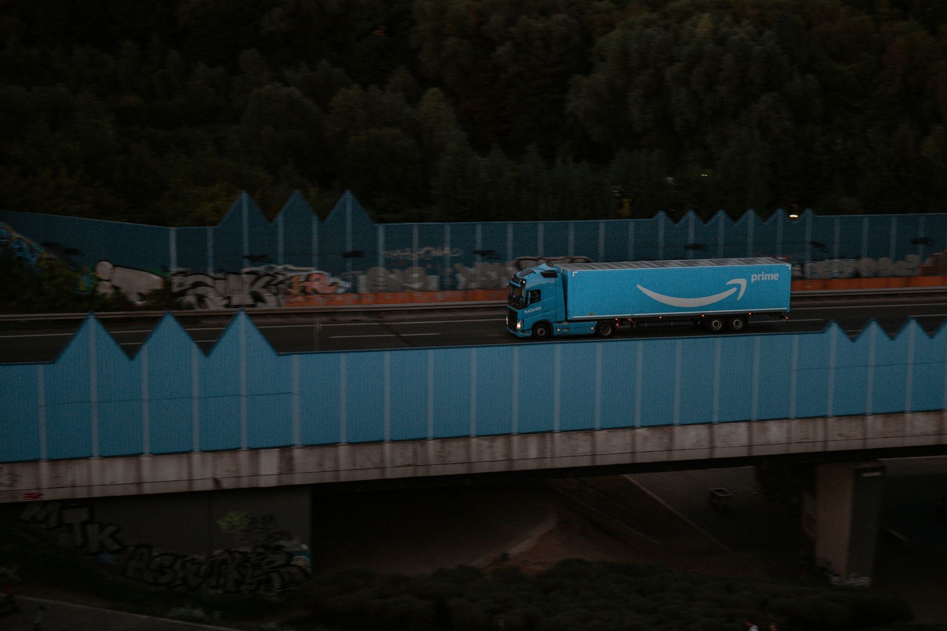 Blue Amazon truck driving on a bridge over a road, with blue sound barriers on each side, at dusk.