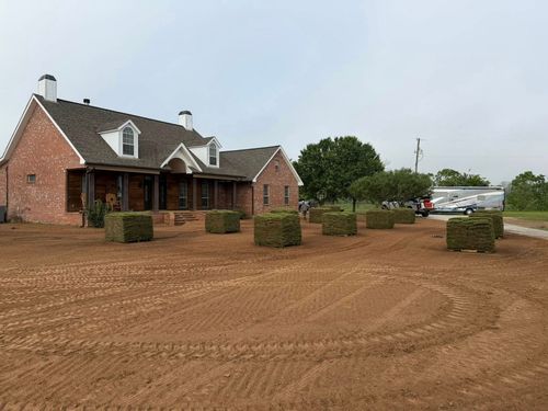 Brick house with a porch and gravel driveway, bordered by trimmed shrubs under a cloudy sky
