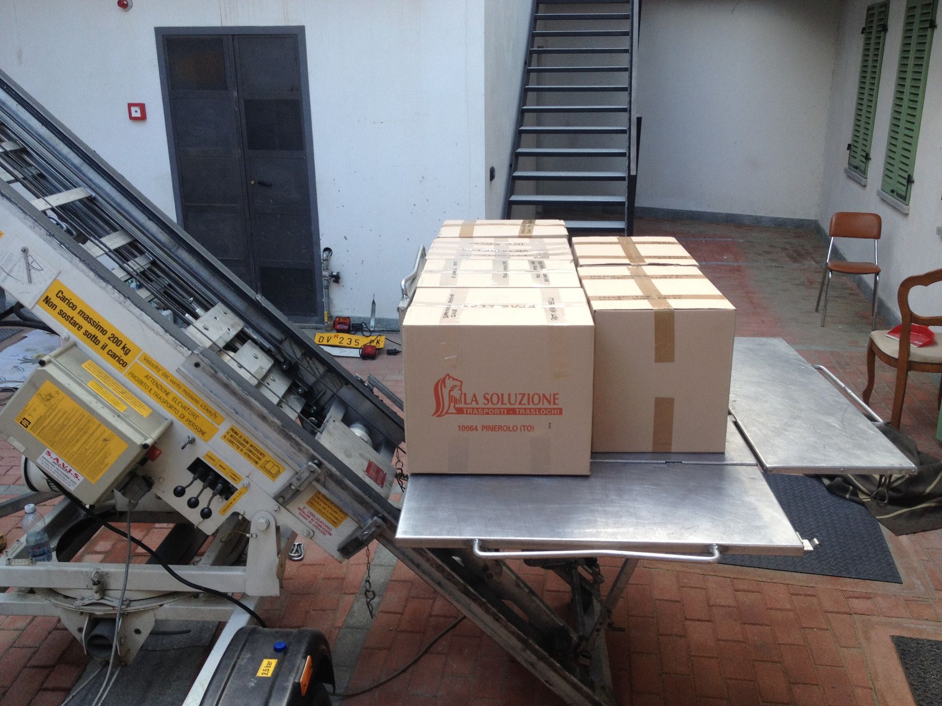 Scatole sulla piattaforma elevatrice, pronte per essere spostate al piano superiore. Porta nera e scale sullo sfondo.