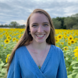 A woman in a blue shirt is smiling in front of a field of sunflowers.