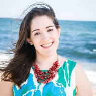 A woman wearing a blue and white dress and a red necklace is smiling on the beach.