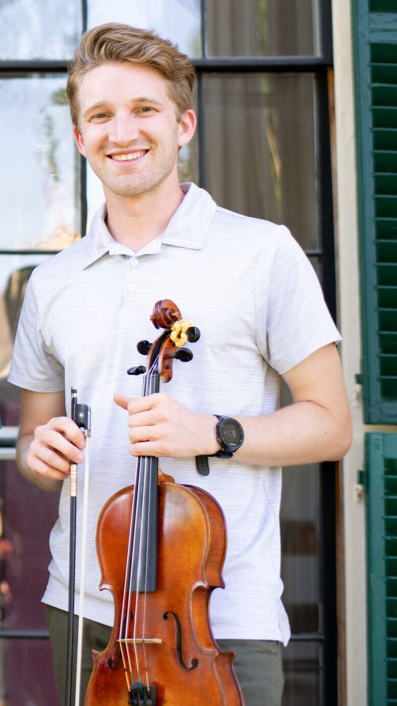 A young man is holding a violin in front of a window.