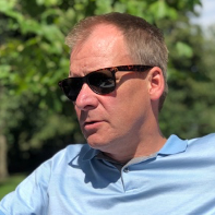 A man wearing sunglasses and a blue shirt is sitting outside.