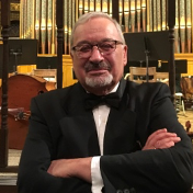 A man in a tuxedo and bow tie is standing with his arms crossed in front of an organ.