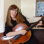 A woman is sitting at a piano holding a cello and smiling.