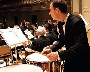 A man in a tuxedo is playing drums in front of an orchestra