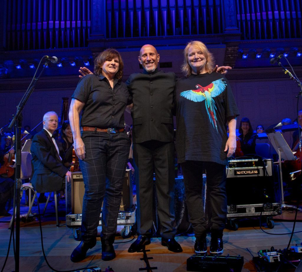 Three people are posing for a picture on a stage with an orchestra in the background