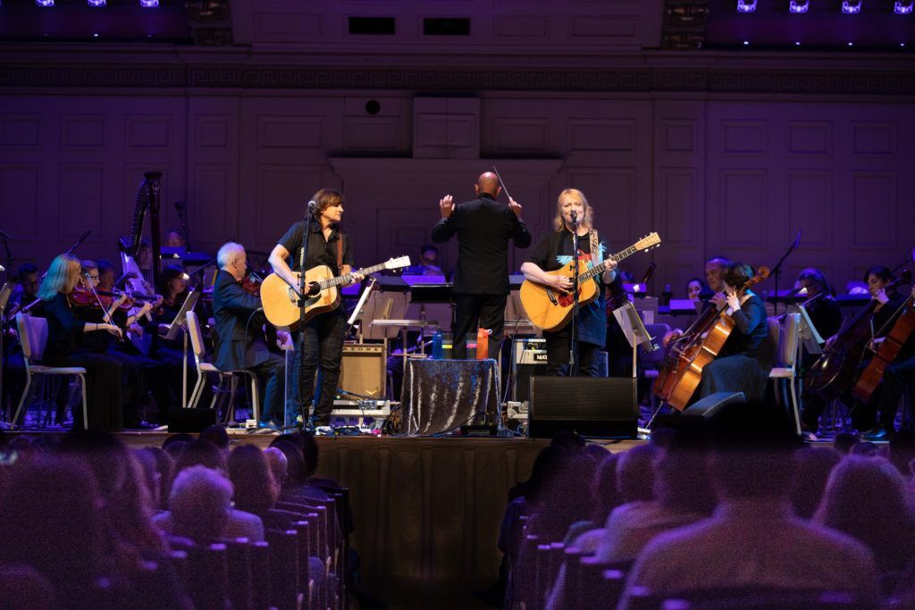 A group of people are playing guitars on a stage in front of an orchestra.