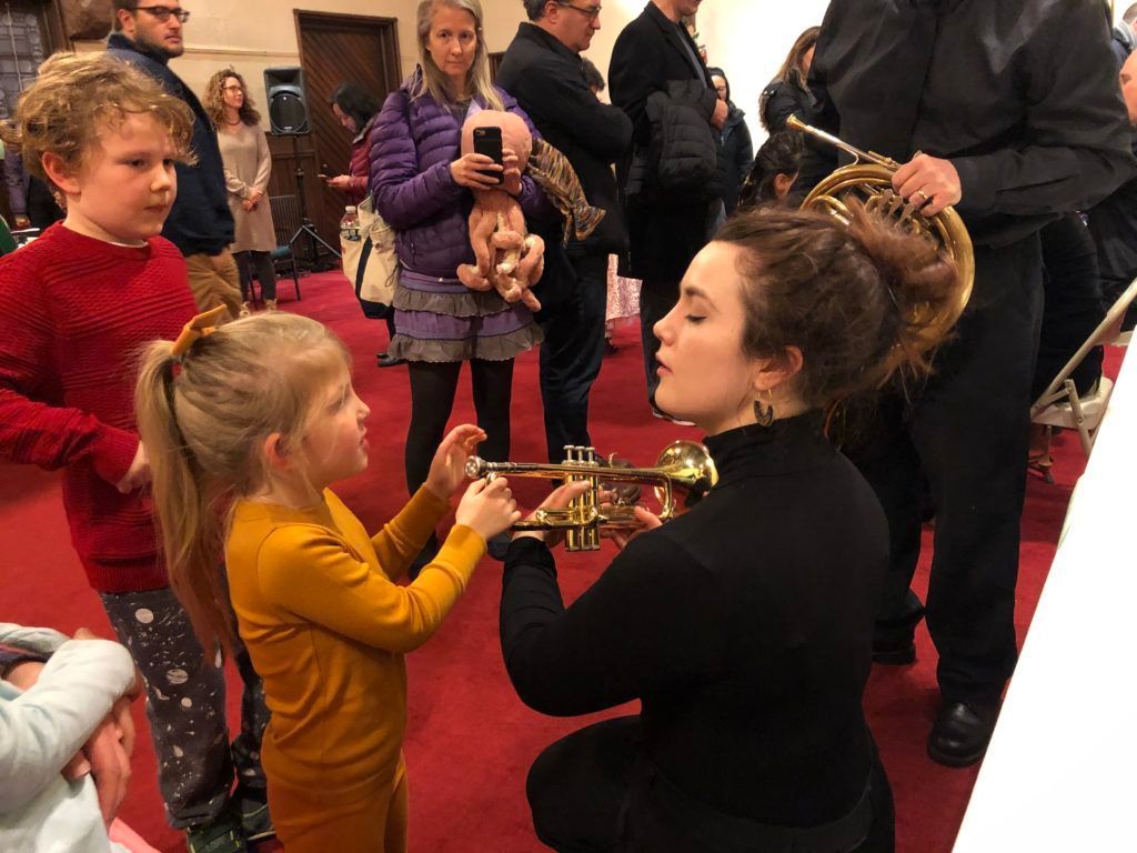 A woman is kneeling down playing a trumpet in front of a group of children.