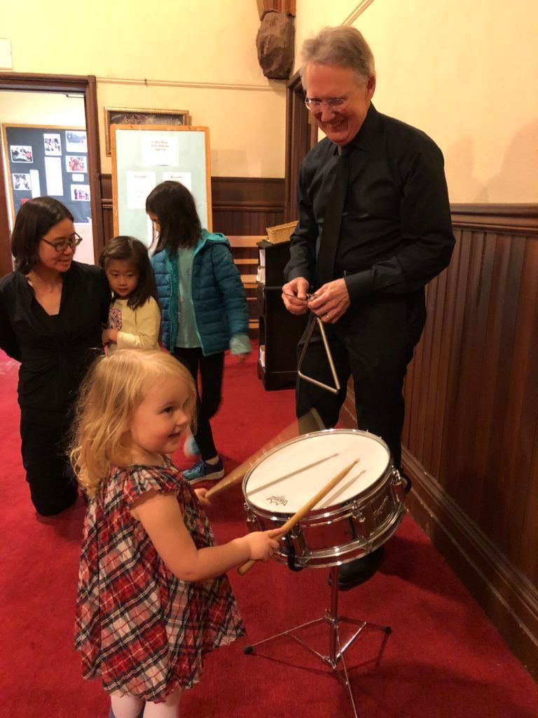 A little girl is playing a drum with a man standing behind her.