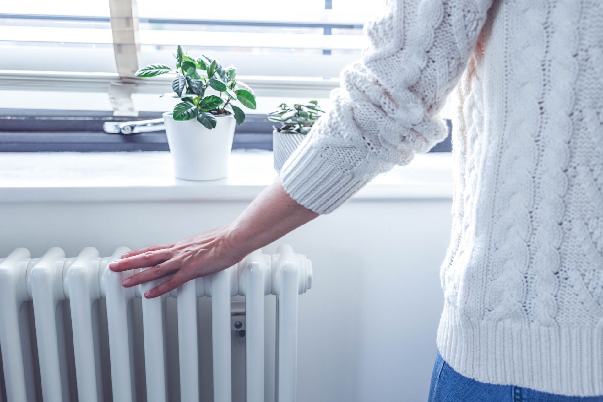 A close-up shot of an unrecognisable woman standing at a sunlit window with her hand on a heating radiator. A close-up shot of an unrecognisable woman standing at a sunlit window with her hand on a heating radiator.