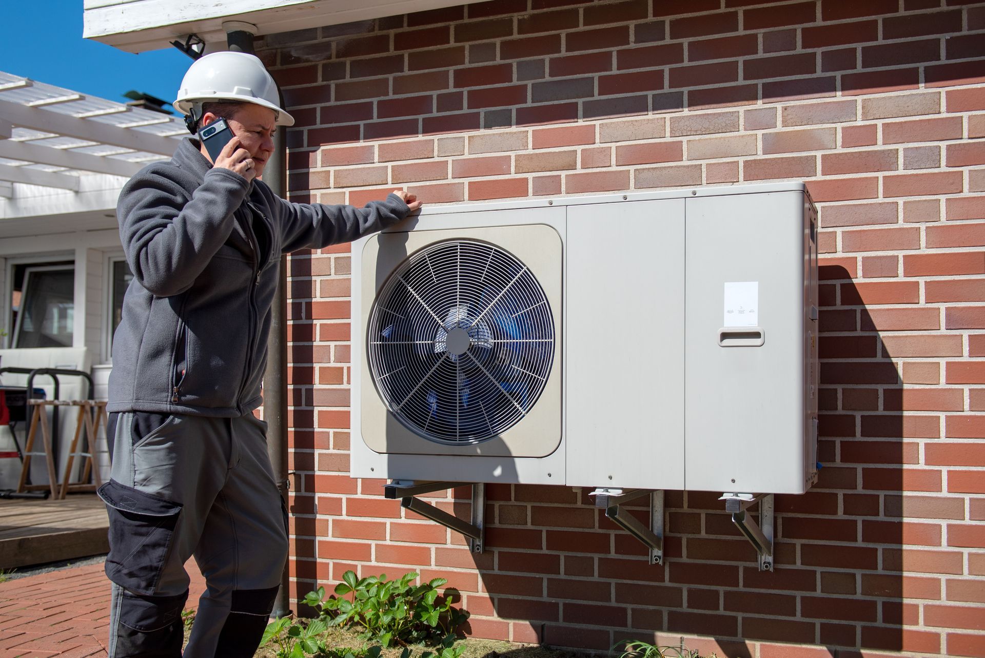 Technician checking outdoor heat pump unit while on phone call in sunlight.