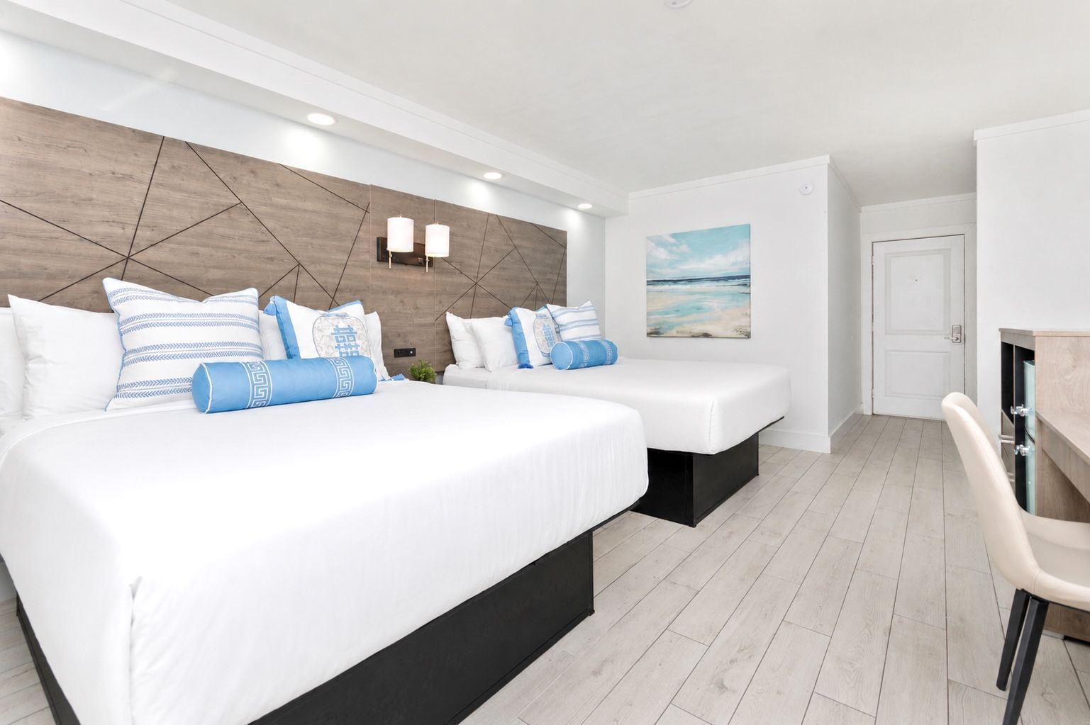 Hotel room with two beds, desk, TV, and mini fridge. Light blue and wood accents.