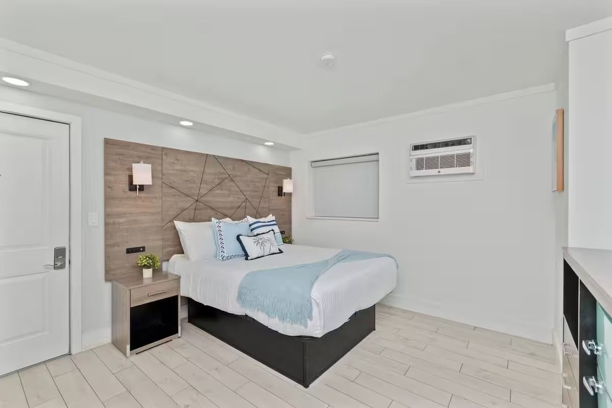 White bedroom with a bed, nightstand, and window. Light blue and white bedding, wooden headboard.