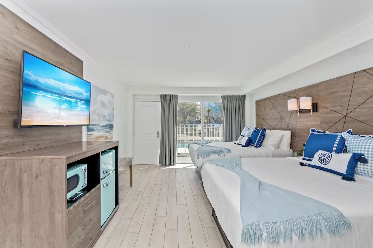 Beach-themed hotel room with two beds, sofa, TV, and mini-fridge. Light blue and white decor. Bright and airy.
