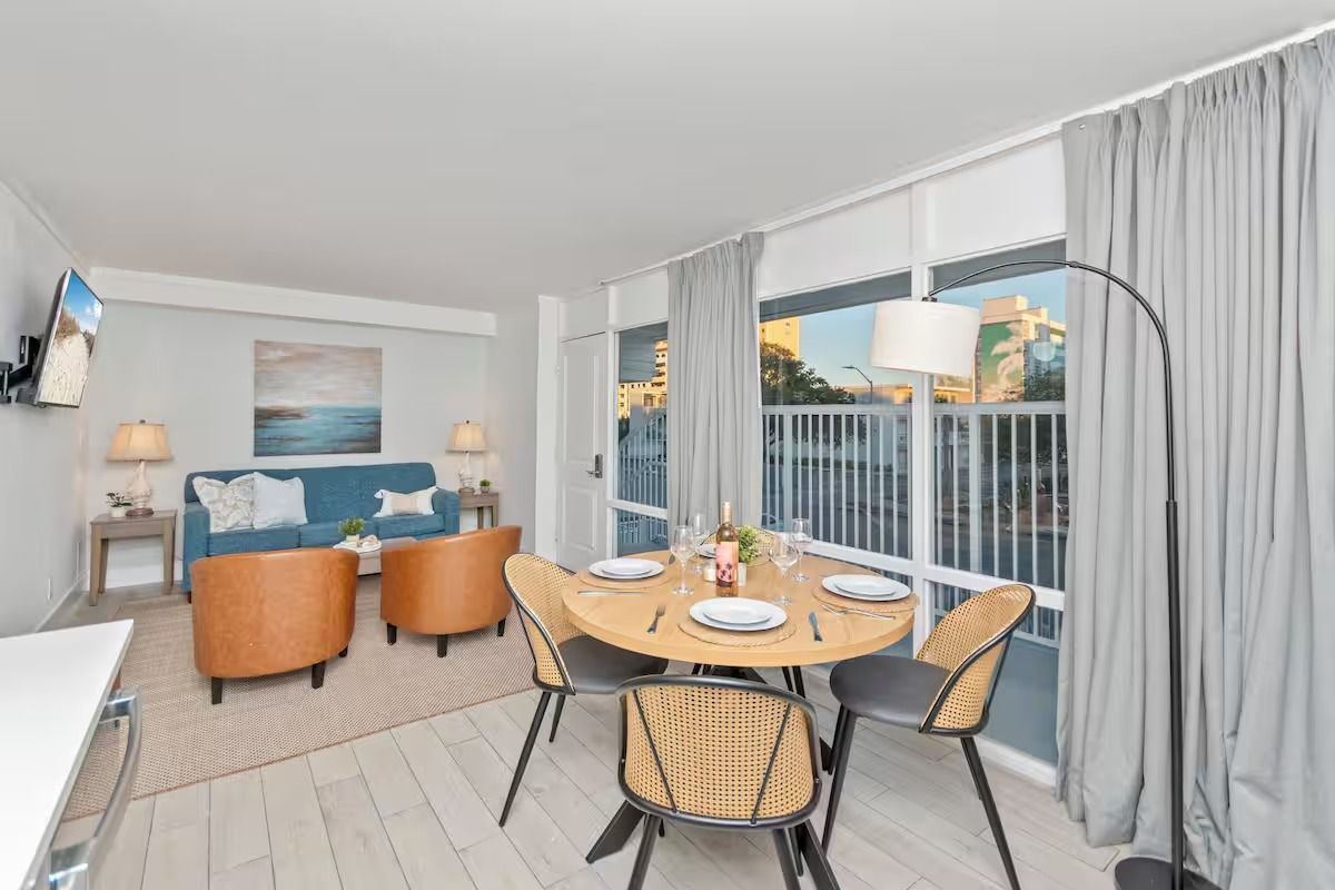 Living area with dining table, sofa, and floor-to-ceiling windows overlooking a balcony and water view.