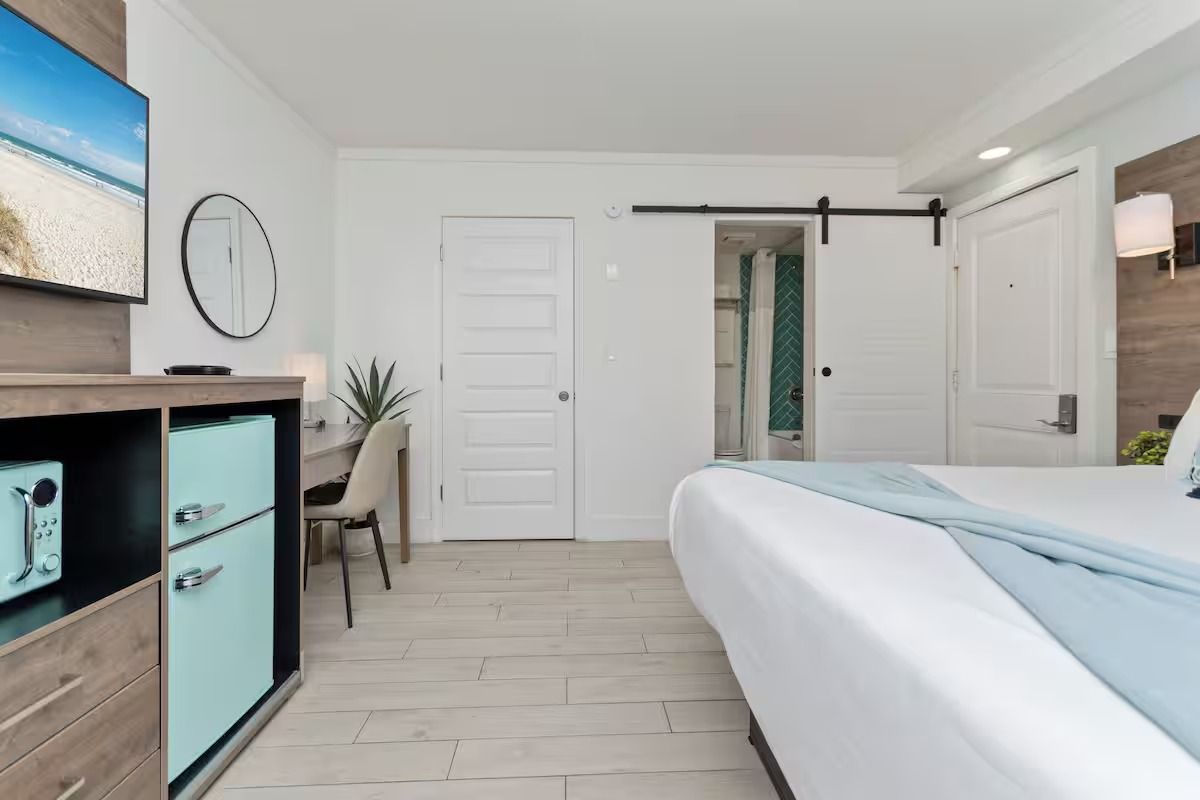 Hotel room with white walls, a turquoise mini-fridge, a desk, a bed, and a sliding barn door leading to the bathroom.