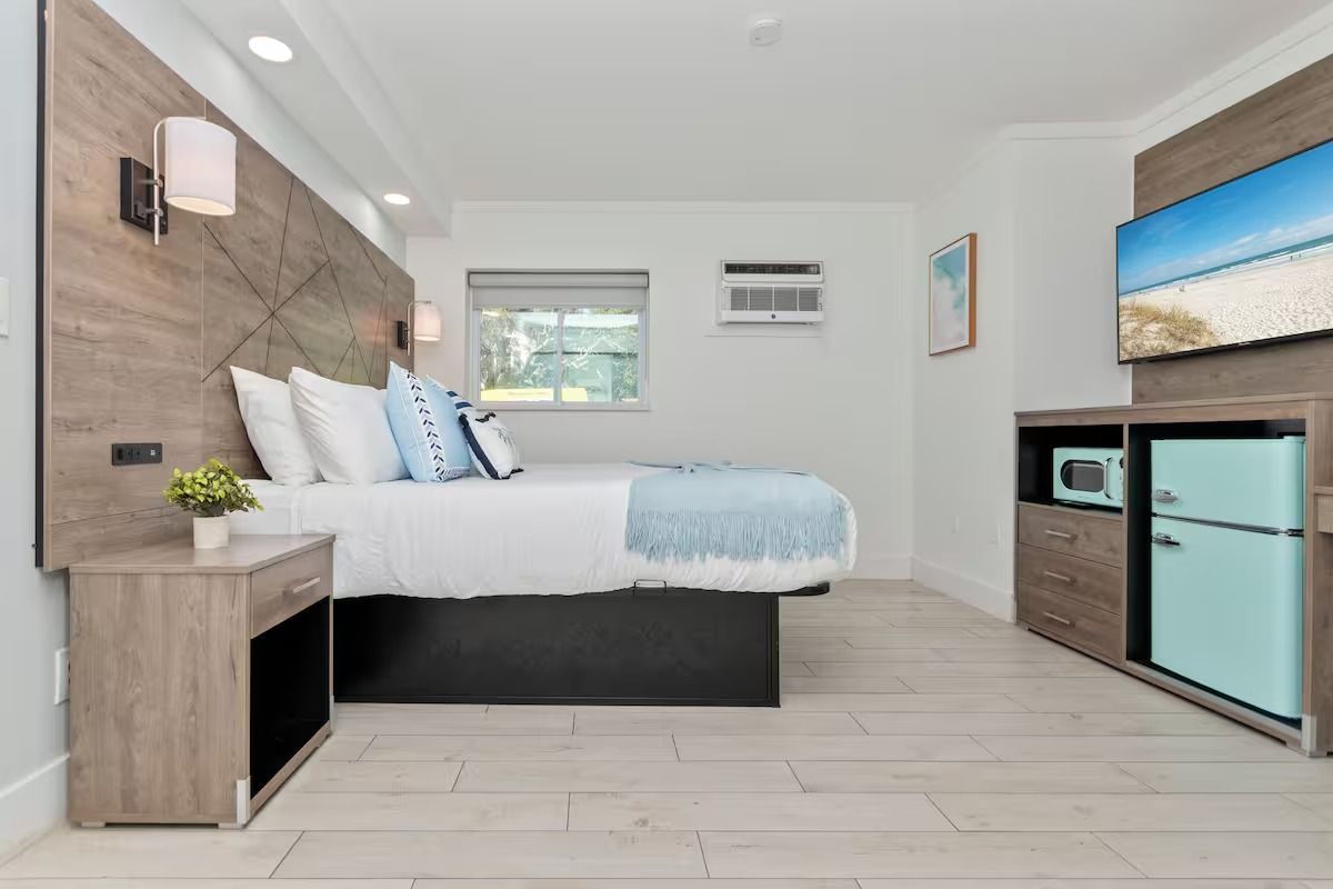 Bedroom with bed, TV, refrigerator, and window. Light wood and blue accents.