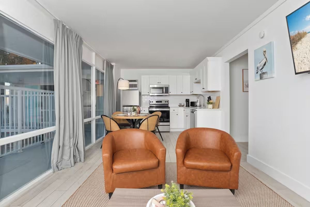Two brown leather armchairs face a kitchen and dining area in a bright, modern room.