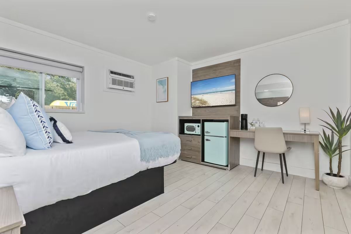 Bedroom with a bed, desk, TV, refrigerator, and window. White walls, wood floor, and neutral decor.