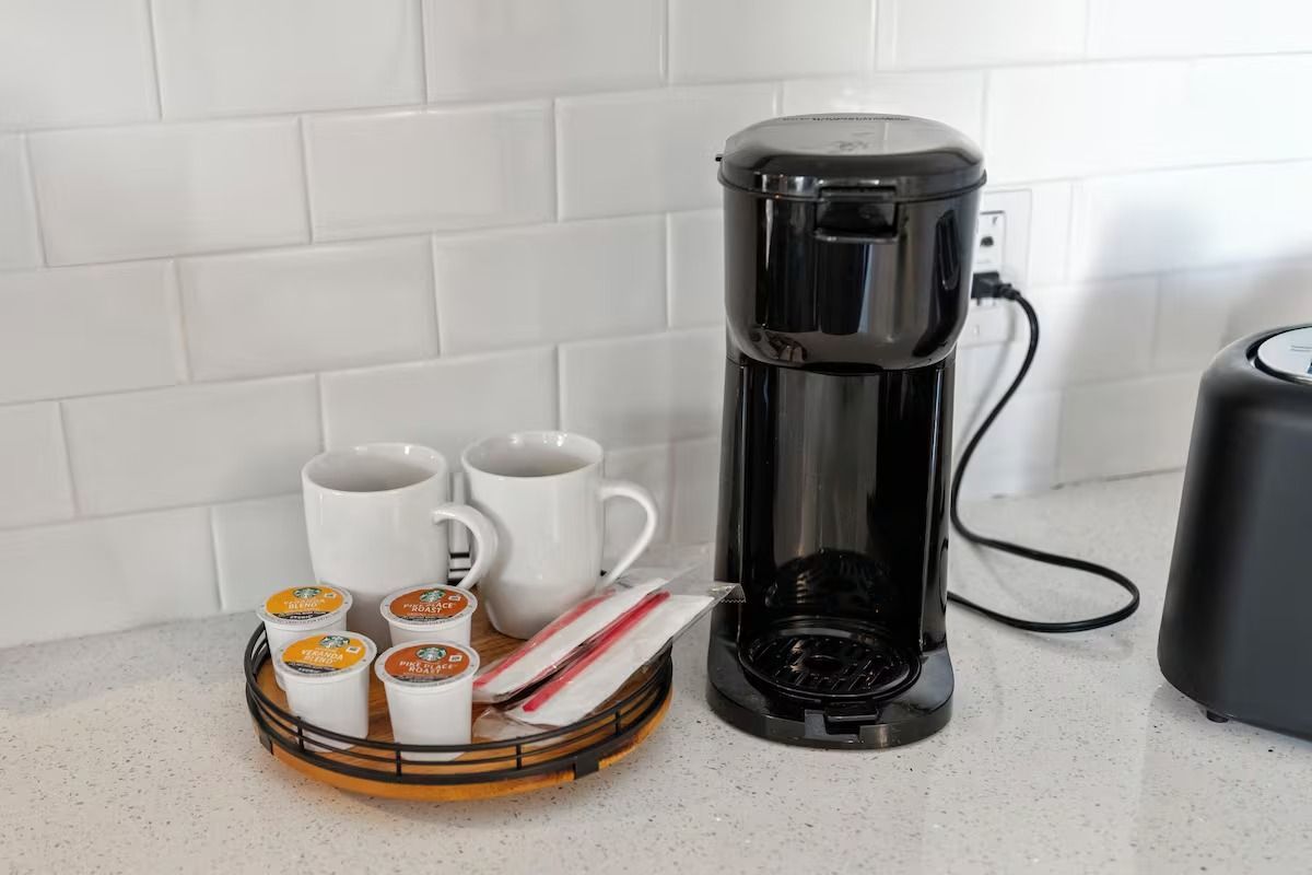 Coffee station on a white counter. Black coffee maker, mugs, K-Cups, sugar packets, and a wooden tray.