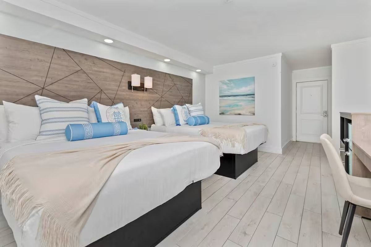Hotel room with two beds, white bedding, light wood accent wall, art, and a desk.
