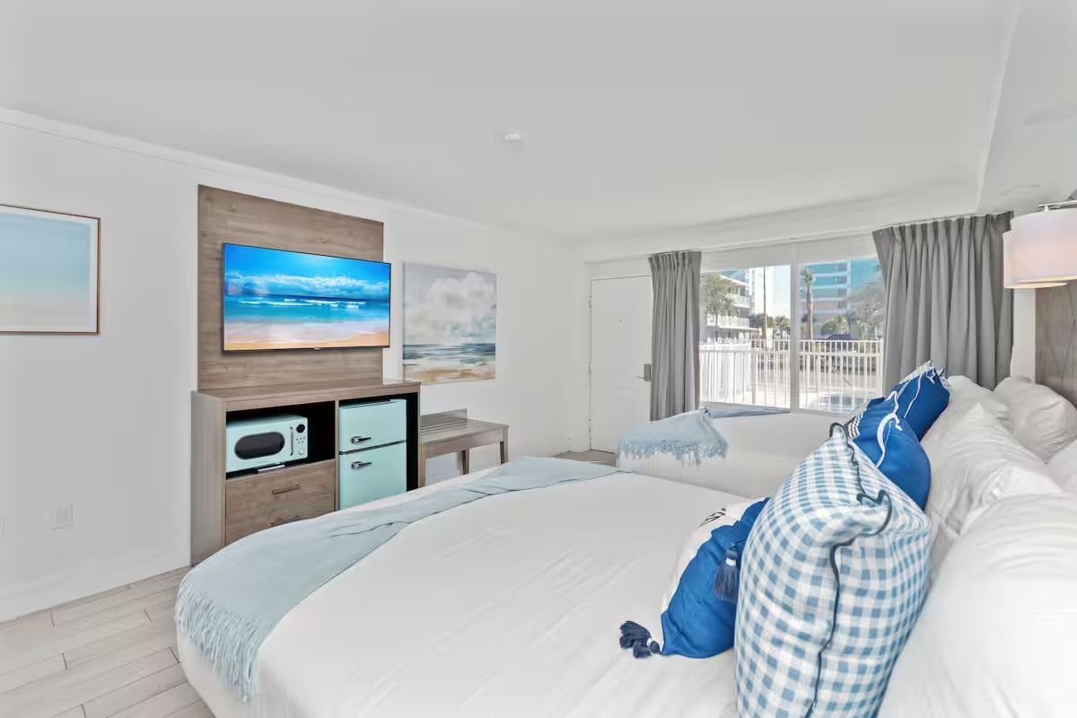 Bedroom with white walls, TV, bed with blue accents, and a mini fridge.