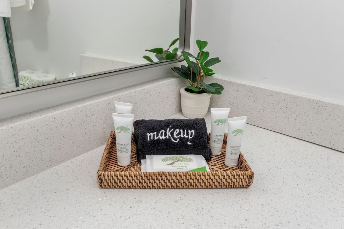 Bathroom toiletries arranged on a woven tray. Includes tubes of lotion, soap, and a makeup washcloth with a small plant.