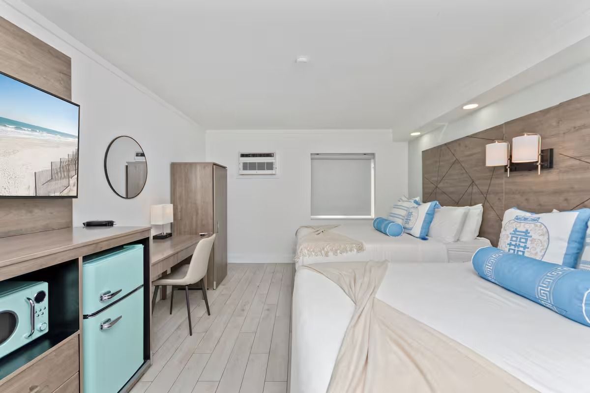Hotel room with two beds, desk, TV, and mini fridge. Light blue and wood accents.