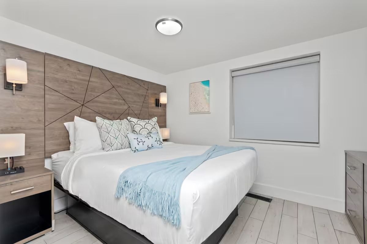 Bedroom with a queen bed, light wood headboard, bedside tables, and a window with a closed blind.