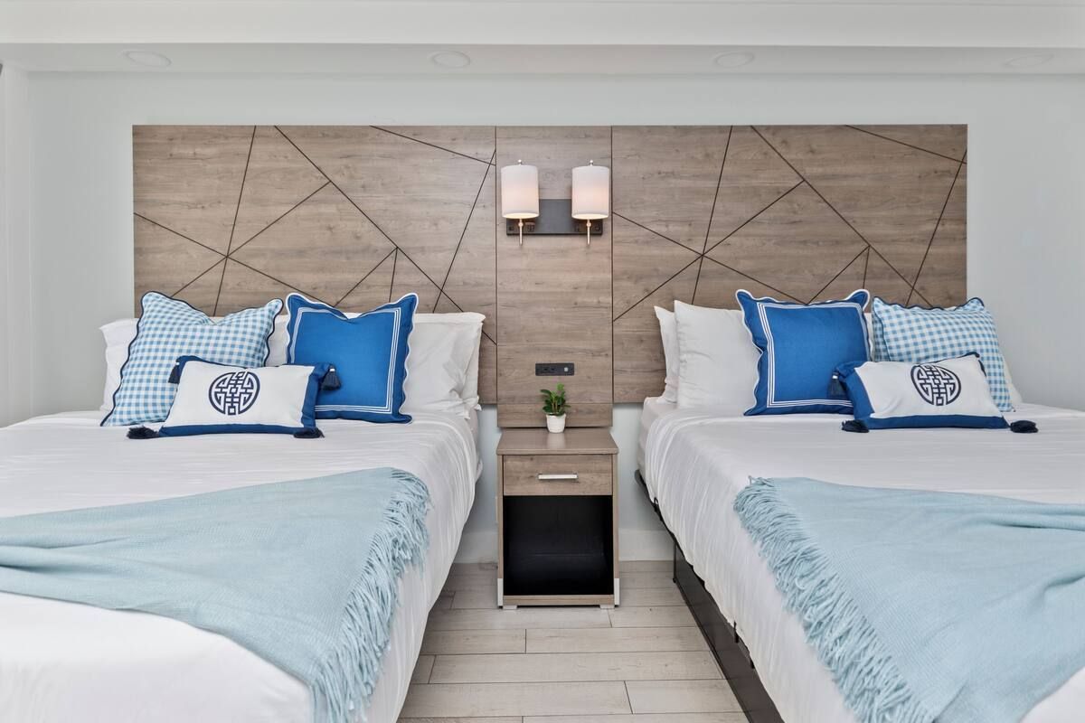 Two beds with blue and white bedding, wooden headboard, small nightstand, and wall sconces.