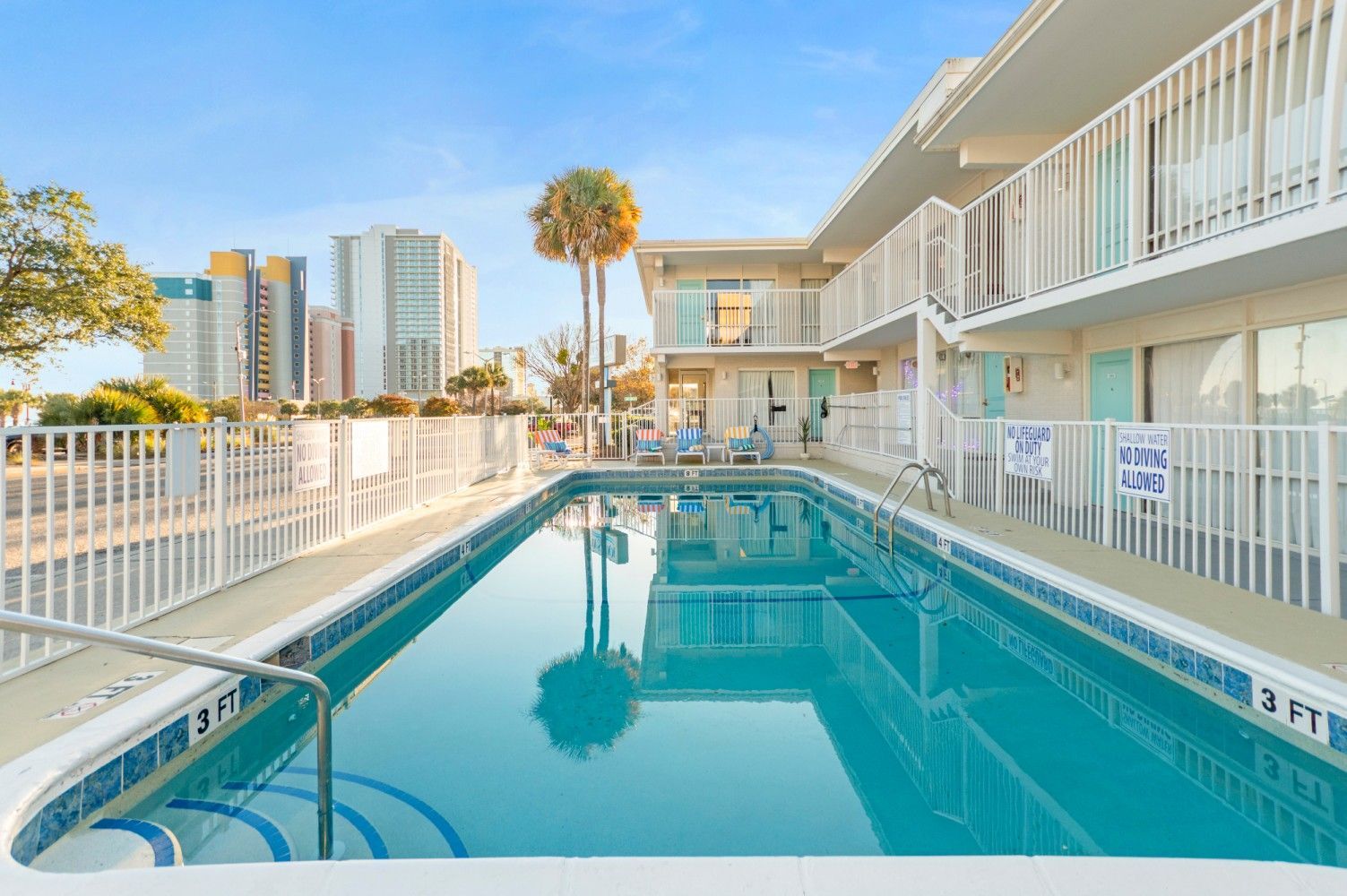Swimming pool in front of a two-story motel. Sunny day, palm tree, and cityscape in the background.